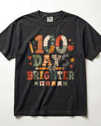 Black Comfort Colors short-sleeve graphic t-shirt laid flat against a white background. The front features a colorful patchwork-style graphic reading '100 DAYS BRIGHTER' with school-themed illustrations like stars, hearts, and pencils.