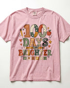 Blossom Comfort Colors short-sleeve crewneck t-shirt featuring a colorful graphic print on the front that reads '100 DAYS BRIGHTER' in patchwork-style letters with autumn motifs like stars, hearts, and an apple.
