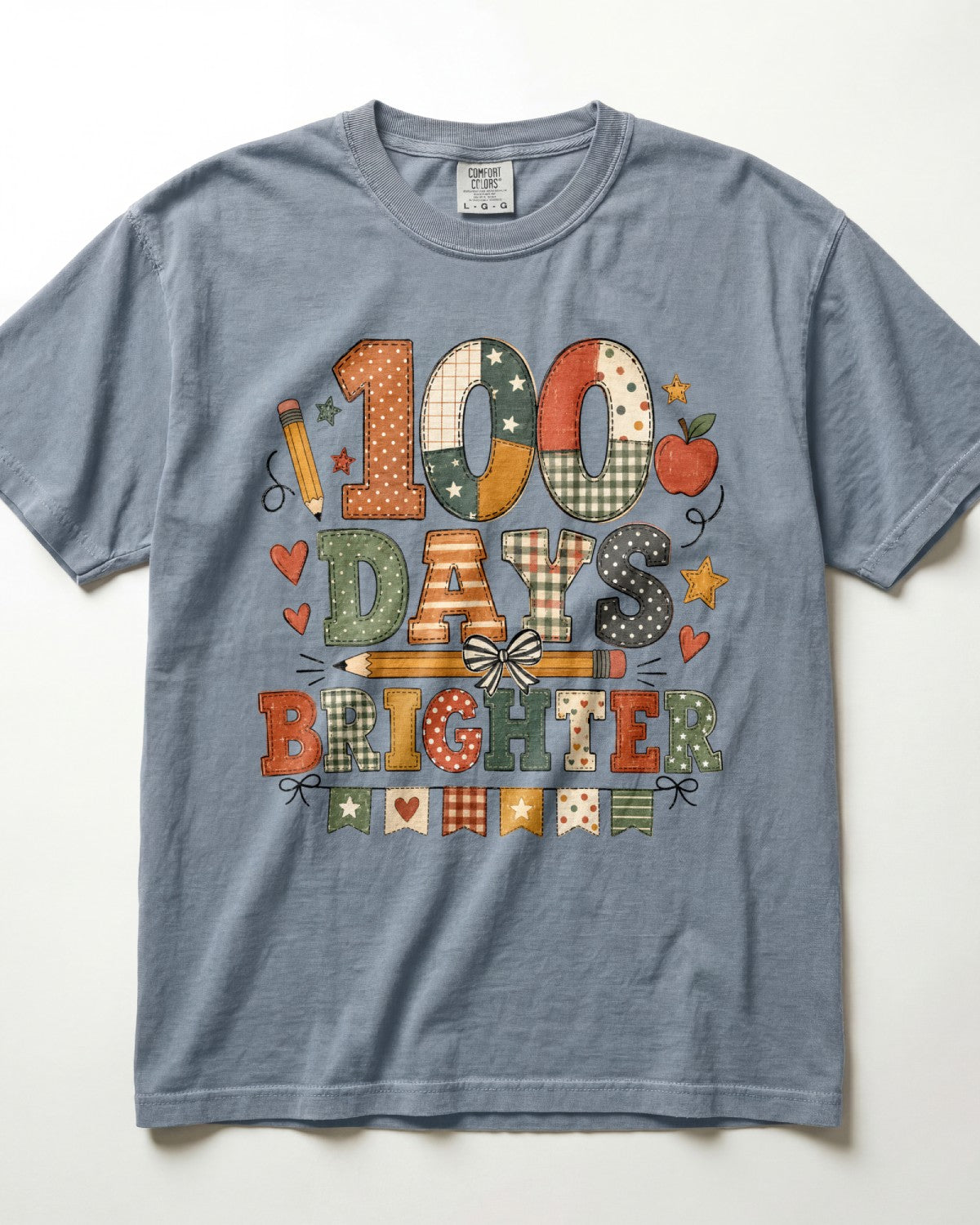 Blue jean Comfort Colors short-sleeve t-shirt with a colorful graphic on the front that reads '100 DAYS BRIGHTER' in patchwork-style letters, decorated with school-themed motifs like stars, hearts, pencils, and an apple, shown on a white background.