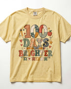 Butter Comfort Colors short-sleeve t-shirt with a colorful graphic on the front that reads '100 DAYS BRIGHTER' in patchwork-style lettering with school-themed motifs like stars, hearts, and an apple, shown flat against a white background.