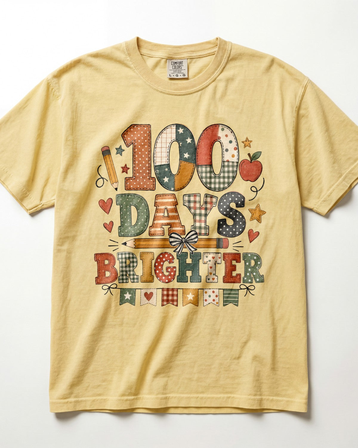 Butter Comfort Colors short-sleeve t-shirt with a colorful graphic on the front that reads '100 DAYS BRIGHTER' in patchwork-style lettering with school-themed motifs like stars, hearts, and an apple, shown flat against a white background.