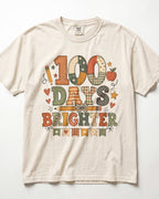 Ivory Comfort Colors short-sleeve t-shirt with a colorful graphic on the front that reads '100 DAYS BRIGHTER' in patchwork-style letters, decorated with stars, hearts, pencils, and an apple.