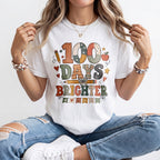 A woman wearing a white graphic t-shirt with a colorful '100 Days Brighter' design, paired with distressed blue jeans and white sneakers, sitting against a plain white background.