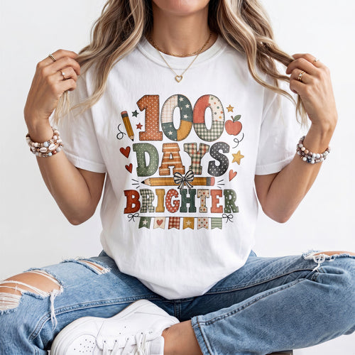 A woman wearing a white graphic t-shirt with a colorful '100 Days Brighter' design, paired with distressed blue jeans and white sneakers, sitting against a plain white background.
