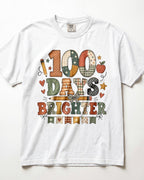 White Comfort Colors short-sleeve crewneck t-shirt featuring a colorful, patchwork-style graphic that reads '100 DAYS BRIGHTER' with school-themed illustrations like pencils, stars, hearts, and an apple.