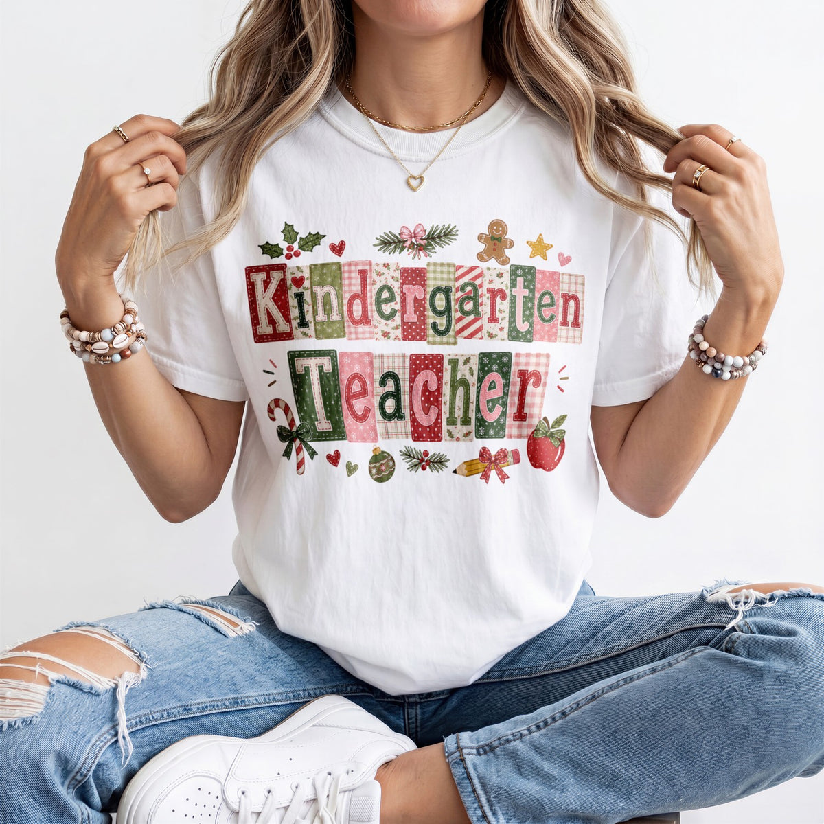 A woman wearing a white graphic t-shirt that reads 'Kindergarten Teacher' in a patchwork pattern with holiday-themed illustrations like candy canes, pinecones, and an apple. She is wearing distressed blue jeans and white sneakers, sitting against a plain white background.