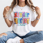 A woman wearing a white graphic t-shirt with a colorful 'Kinder Garden Crew' text design, paired with distressed blue jeans and white sneakers, sitting cross-legged against a plain white background.
