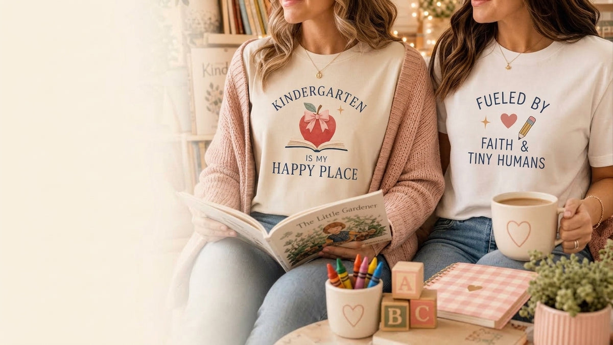 Two kindergarten teachers wearing graphic tees, "Kindergarten Is My Happy Place" and "Fueled by Faith & Tiny Humans," surrounded by classroom books and supplies