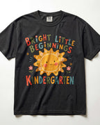 Black Comfort Colors kindergarten teacher shirt featuring a colorful sun graphic and “Bright Little Beginnings” text, displayed in a flat lay.