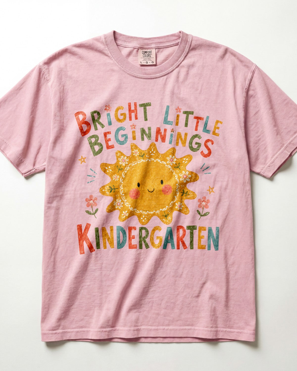 Blossom Comfort Colors kindergarten teacher shirt featuring a colorful sun graphic and “Bright Little Beginnings” text, displayed in a flat lay.