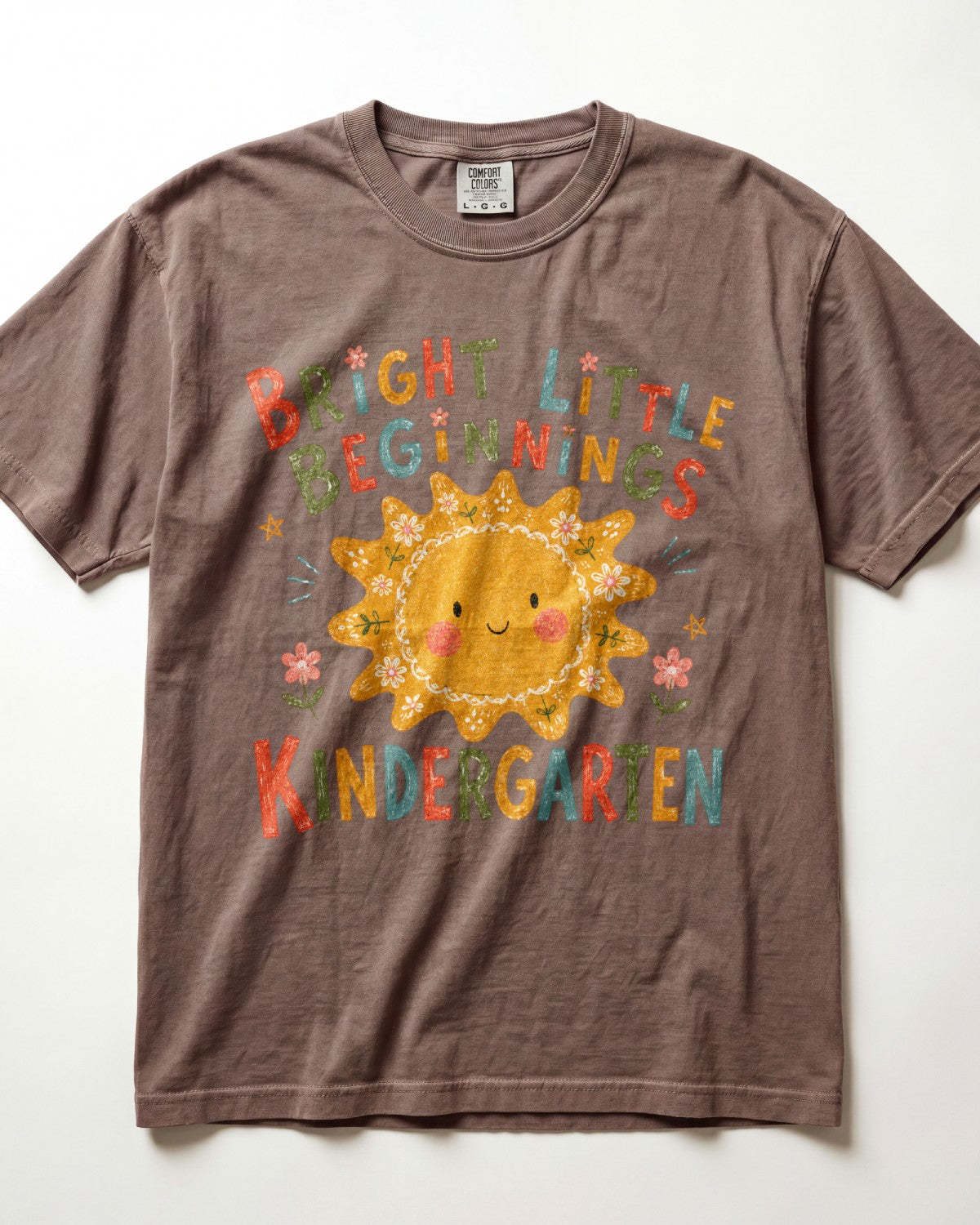 Espresso Comfort Colors kindergarten teacher shirt featuring a colorful sun graphic and “Bright Little Beginnings” text, displayed in a flat lay.