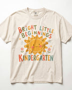 Ivory Comfort Colors kindergarten teacher shirt featuring a colorful sun graphic and “Bright Little Beginnings” text, displayed in a flat lay.