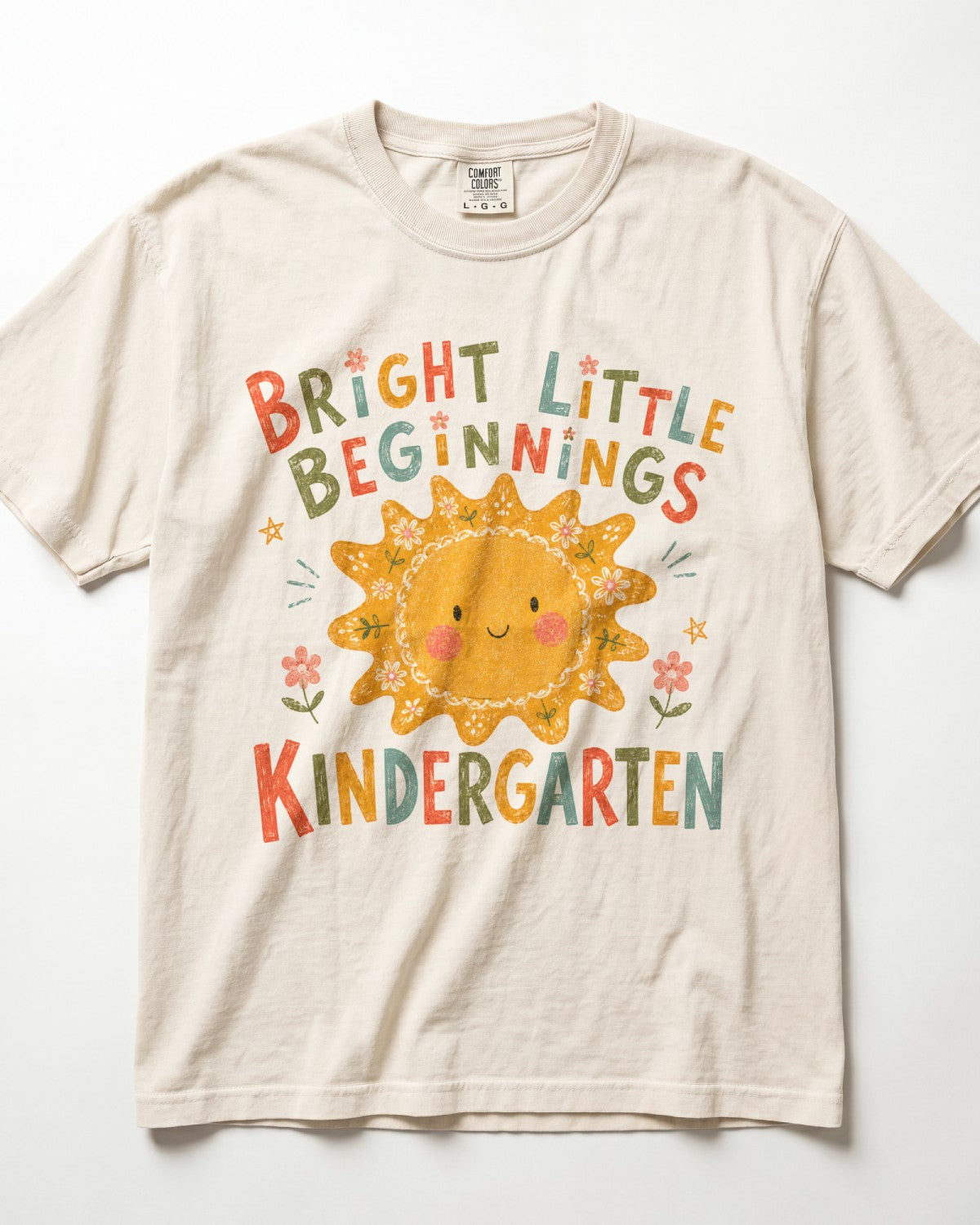 Ivory Comfort Colors kindergarten teacher shirt featuring a colorful sun graphic and “Bright Little Beginnings” text, displayed in a flat lay.