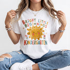 Woman wearing a white Comfort Colors kindergarten teacher shirt featuring a colorful sun graphic and “Bright Little Beginnings” text.