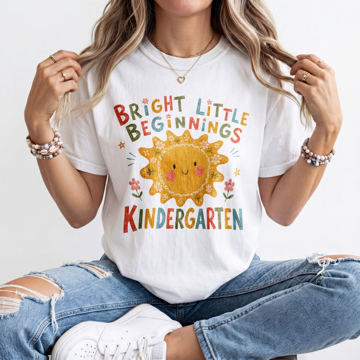 Woman wearing a white Comfort Colors kindergarten teacher shirt featuring a colorful sun graphic and “Bright Little Beginnings” text.