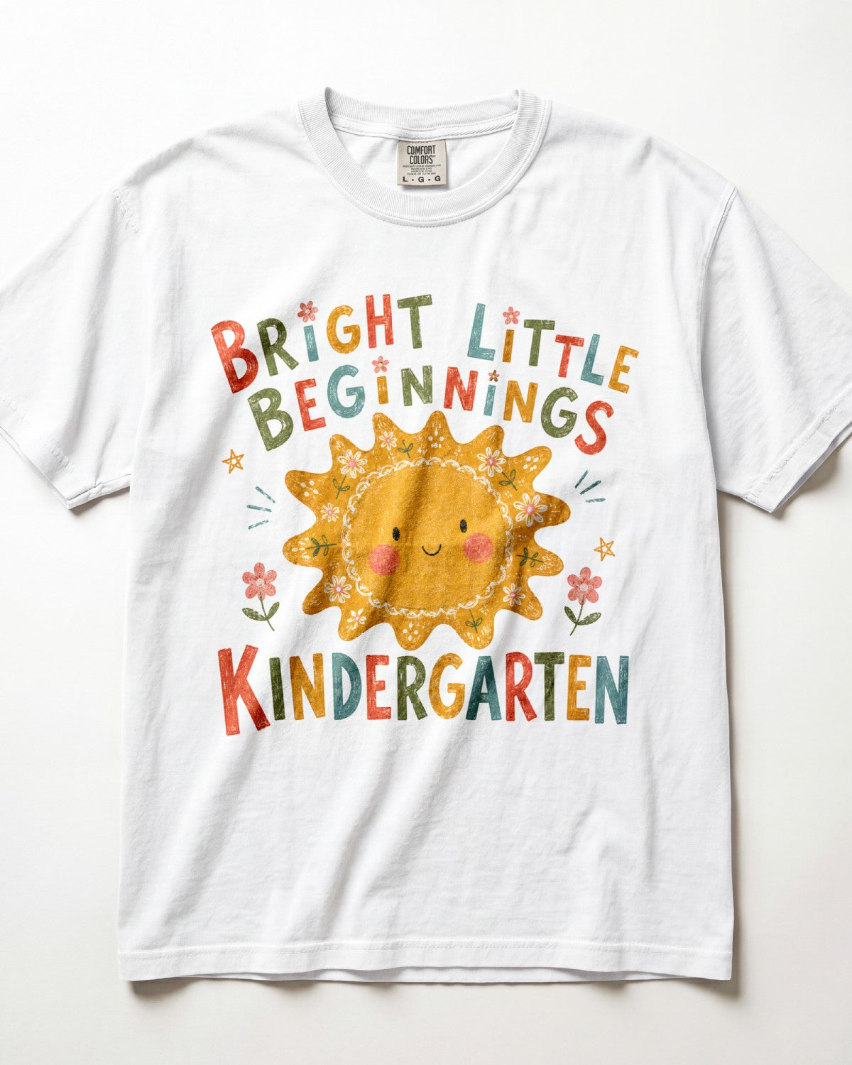 White Comfort Colors kindergarten teacher shirt featuring a colorful sun graphic and “Bright Little Beginnings” text, displayed in a flat lay.