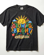 A black short-sleeve t-shirt with a colorful graphic on the front that reads 'GROWING BRIGHT IN KINDERGARTEN' with a smiling sun, clouds, stars, and school-themed icons.