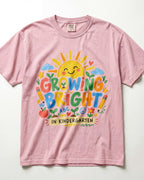 A light pink/blossom short-sleeve t-shirt with a colorful graphic on the front that reads 'GROWING BRIGHT IN KINDERGARTEN' surrounded by playful school-themed illustrations like a smiling sun, clouds, pencils, an apple, and flowers.