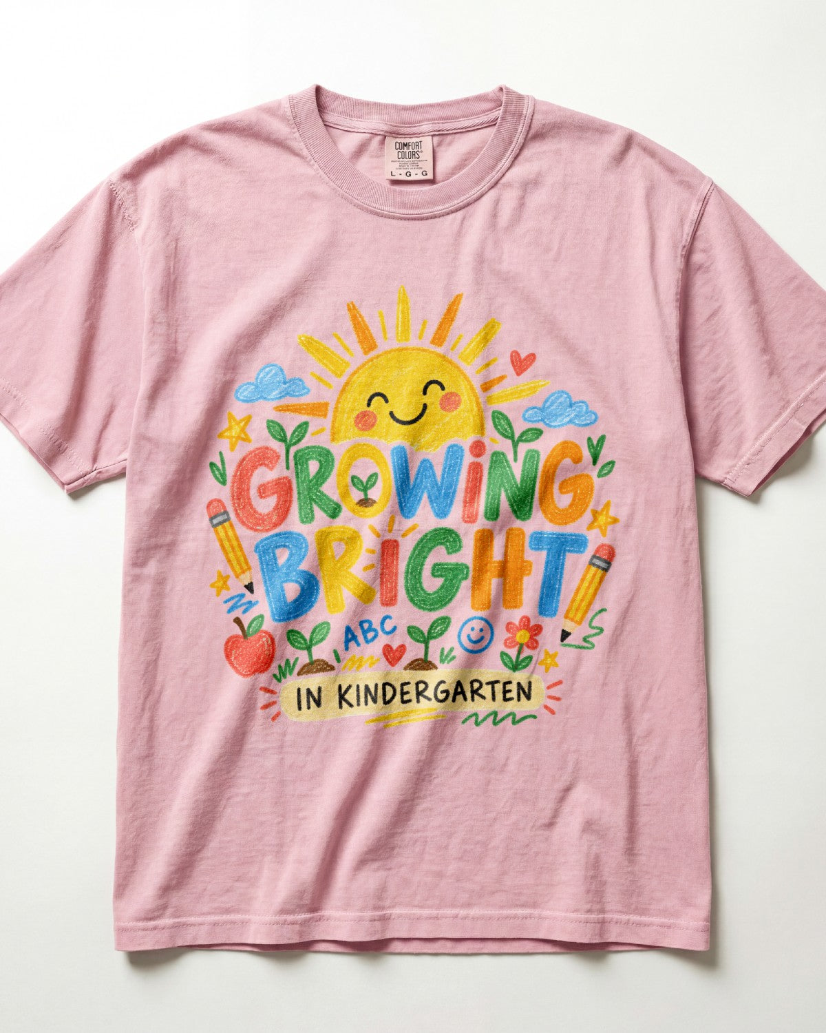 A light pink/blossom short-sleeve t-shirt with a colorful graphic on the front that reads 'GROWING BRIGHT IN KINDERGARTEN' surrounded by playful school-themed illustrations like a smiling sun, clouds, pencils, an apple, and flowers.