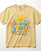 A light yellow/butter short-sleeve t-shirt with a colorful graphic on the front that reads 'GROWING BRIGHT IN KINDERGARTEN' surrounded by playful school-themed illustrations like a smiling sun, clouds, hearts, and pencils.