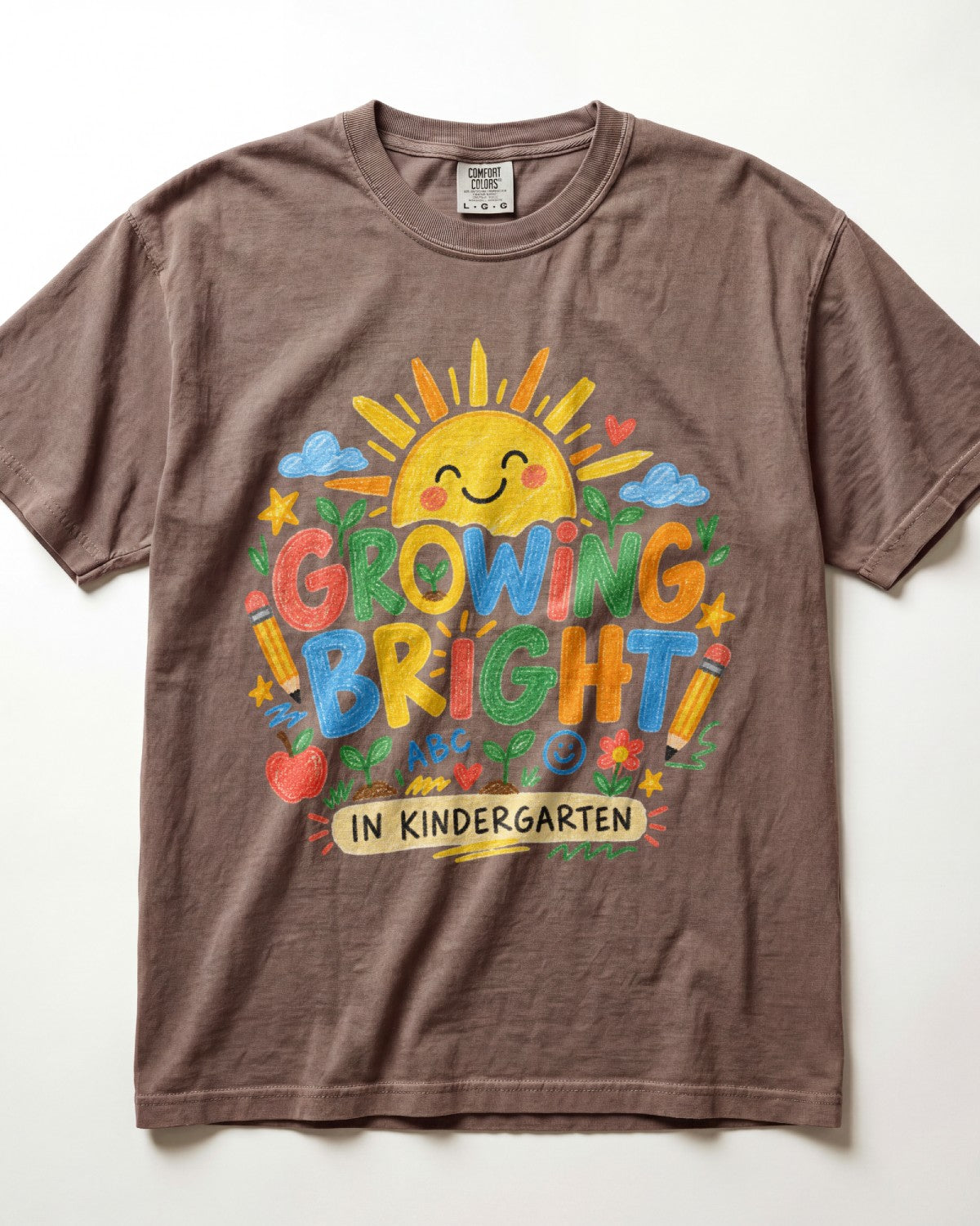 A brown/espresso short-sleeve crewneck t-shirt featuring a colorful graphic on the front with a smiling sun, crayons, stars, and the text 'GROWING BRIGHT! IN KINDERGARTEN'.