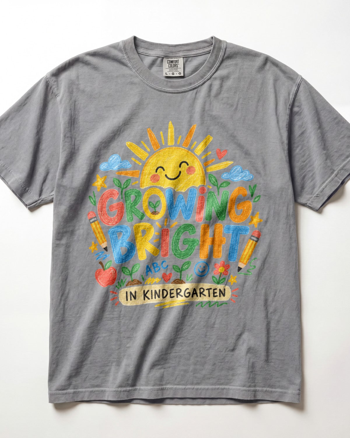 A heather grey short-sleeve t-shirt with a colorful graphic on the front featuring a smiling sun, the words 'GROWING BRIGHT' in playful multicolor letters, and 'IN KINDERGARTEN' on a banner, laid flat against a white background.