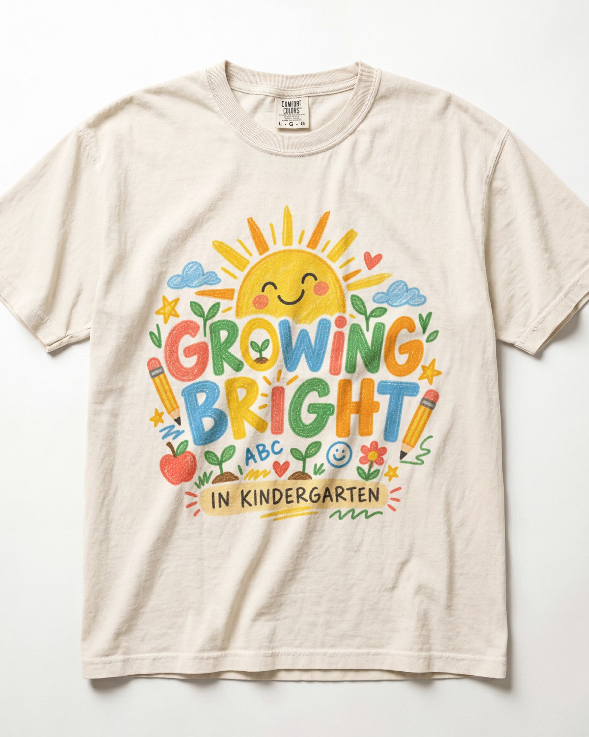 A cream-colored/ivory short-sleeve t-shirt with a colorful graphic on the front that reads 'GROWING BRIGHT IN KINDERGARTEN' surrounded by playful school-themed illustrations like a smiling sun, clouds, pencils, and an apple.