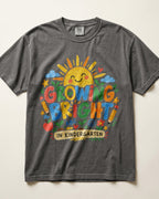 A dark gray/pepper heather t-shirt with a colorful graphic print that reads 'GLOWING BRIGHT IN KINDERGARTEN' featuring a smiling sun, clouds, and school-themed illustrations, laid flat against a light beige background.