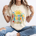 A woman wearing a cream-colored/ivory graphic t-shirt with a colorful 'Growing Bright in Kindergarten' sun design, paired with distressed blue jeans and white sneakers.