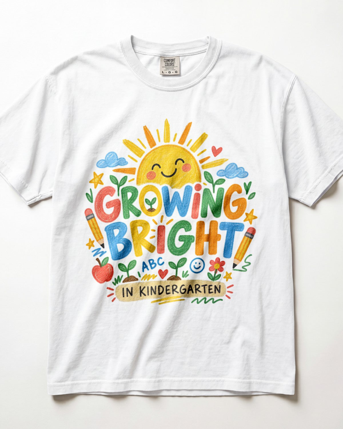 A white short-sleeve crewneck t-shirt featuring a colorful, playful graphic on the front that reads 'GROWING BRIGHT IN KINDERGARTEN' surrounded by illustrations of a smiling sun, clouds, pencils, an apple, and stars.