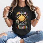 A woman wearing a black graphic t-shirt with a colorful 'HELLO! LITTLE LEARNERS' sun design, paired with distressed blue jeans and white sneakers.
