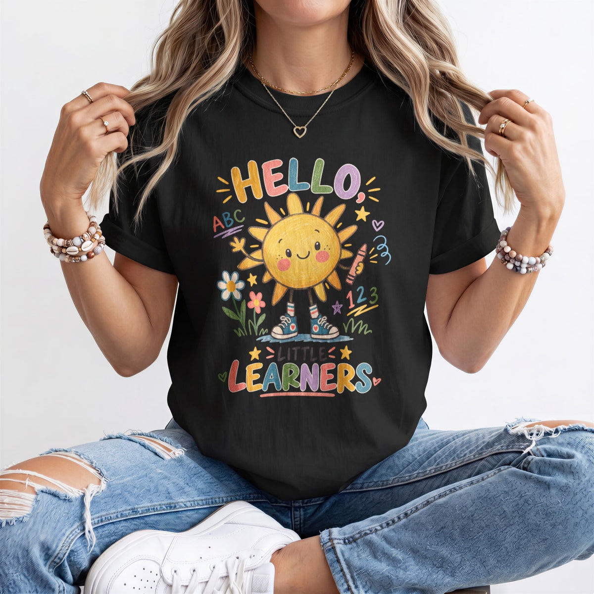 A woman wearing a black graphic t-shirt with a colorful 'HELLO! LITTLE LEARNERS' sun design, paired with distressed blue jeans and white sneakers.