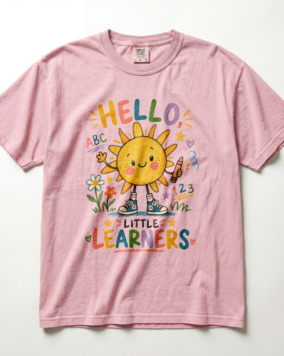 A blossom/pink short-sleeve t-shirt with a colorful cartoon graphic of a smiling sun and the text 'HELLO, LITTLE LEARNERS' surrounded by flowers, stars, and numbers.