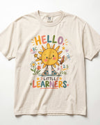 A cream-colored/ivory short-sleeve t-shirt with a colorful graphic of a smiling sun and the text 'HELLO, LITTLE LEARNERS' on the front, displayed flat against a white background.