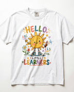 A white short-sleeve crewneck t-shirt featuring a colorful cartoon graphic of a smiling sun with legs, surrounded by flowers, stars, and the text 'HELLO, ABC, 123 LITTLE LEARNERS'.