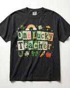 A black short-sleeve crewneck t-shirt featuring a colorful graphic that reads 'One Lucky Teacher' in St. Patrick's Day themed tiles, decorated with shamrocks, a rainbow, a pot of gold, a horseshoe, and an apple, laid flat against a white background.