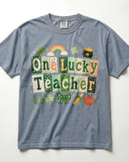A heathered slate blue/blue jean short-sleeve t-shirt featuring a St. Patrick's Day graphic that reads 'One Lucky Teacher' in patchwork-style green and beige letters, decorated with a rainbow, clover, shamrock, pot of gold, horseshoe, bow, pencil, and apple on a white background.