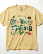 A pale yellow/butter short-sleeve t-shirt with a colorful St. Patrick's Day graphic that reads 'One Lucky Teacher' in patchwork-style letters, decorated with a rainbow, shamrock, pot of gold, horseshoe, bow, pencil, and apple.
