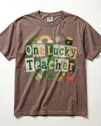 A brown/espresso short-sleeve crewneck t-shirt featuring a St. Patrick's Day graphic that reads 'One Lucky Teacher' in a patchwork pattern of green, white, and orange squares, decorated with a rainbow, shamrocks, a pot of gold, and a carrot.