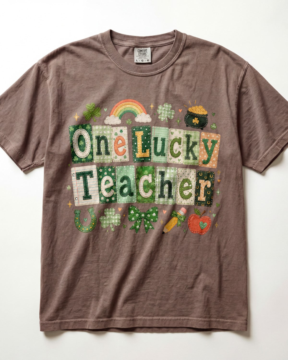 A brown/espresso short-sleeve crewneck t-shirt featuring a St. Patrick's Day graphic that reads 'One Lucky Teacher' in a patchwork pattern of green, white, and orange squares, decorated with a rainbow, shamrocks, a pot of gold, and a carrot.