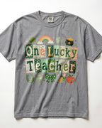 A gray short-sleeve crewneck t-shirt featuring a St. Patrick's Day graphic that reads 'One Lucky Teacher' in patchwork-style letters with green, orange, and white patterns.