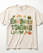 A cream-colored/ivory short-sleeve t-shirt featuring a colorful St. Patrick's Day graphic that reads 'One Lucky Teacher' in patchwork-style letters, decorated with a rainbow, shamrocks, a pot of gold, a horseshoe, a bow, a carrot, and an apple.