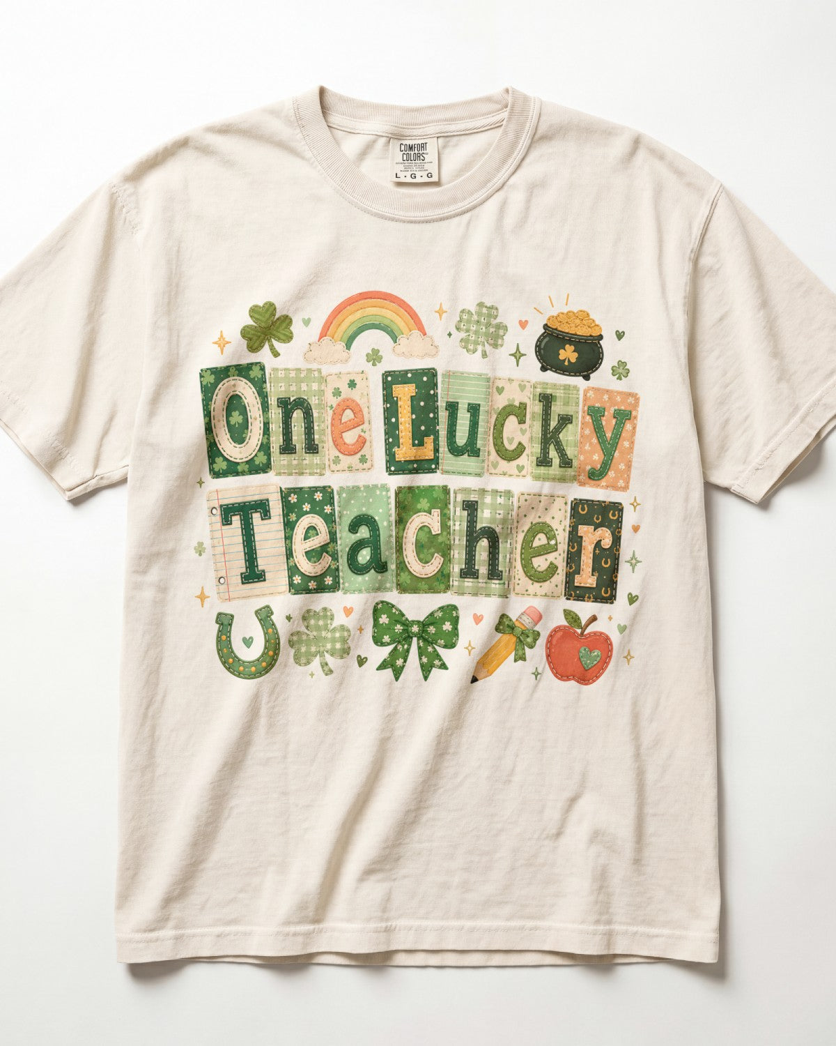 A cream-colored/ivory short-sleeve t-shirt featuring a colorful St. Patrick's Day graphic that reads 'One Lucky Teacher' in patchwork-style letters, decorated with a rainbow, shamrocks, a pot of gold, a horseshoe, a bow, a carrot, and an apple.
