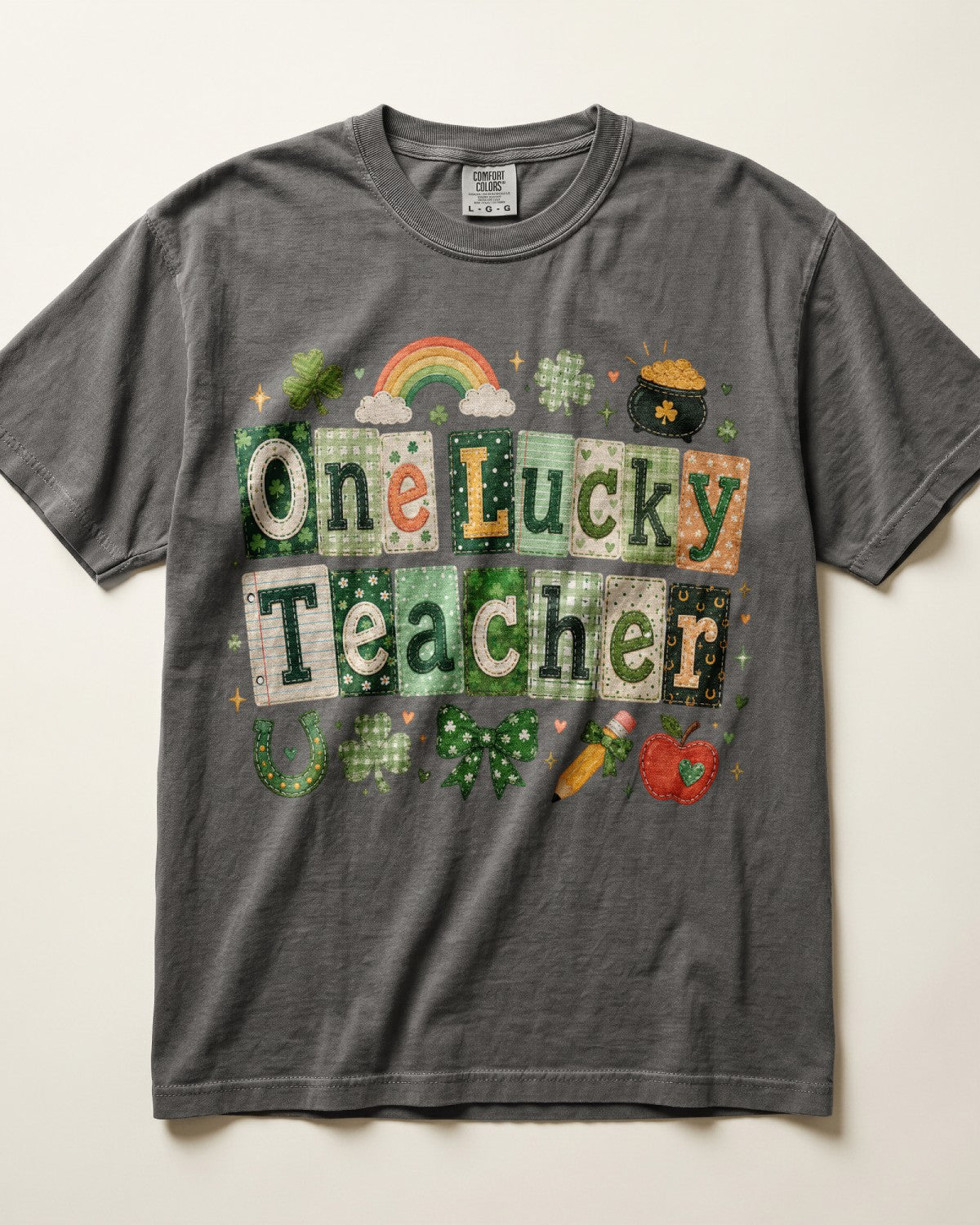 A dark gray/pepper crewneck t-shirt featuring a colorful 'One Lucky Teacher' graphic with St. Patrick's Day themed illustrations like a rainbow, leprechaun pot, and shamrocks, laid flat against a light beige background.