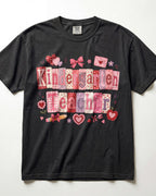 A black short-sleeve t-shirt with a 'Kindergarten Teacher' graphic made of pink and red patterned tiles, decorated with hearts, bows, and flowers, laid flat against a white background.