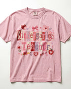 A light pink/blossom short-sleeve crewneck t-shirt featuring a colorful graphic on the front that reads 'Kindergarten Teacher' in patchwork-style letters, decorated with red hearts, bows, an envelope, and small school-themed icons.