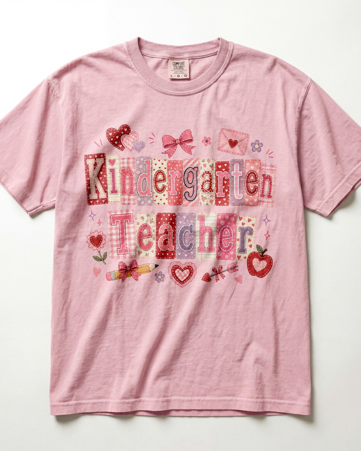 A light pink/blossom short-sleeve crewneck t-shirt featuring a colorful graphic on the front that reads 'Kindergarten Teacher' in patchwork-style letters, decorated with red hearts, bows, an envelope, and small school-themed icons.