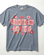 A slate blue/blue jean short-sleeve t-shirt with a 'Kindergarten Teacher' graphic on the front, featuring a patchwork-style design in pink, red, and white with hearts, bows, and strawberries, laid flat against a white background.