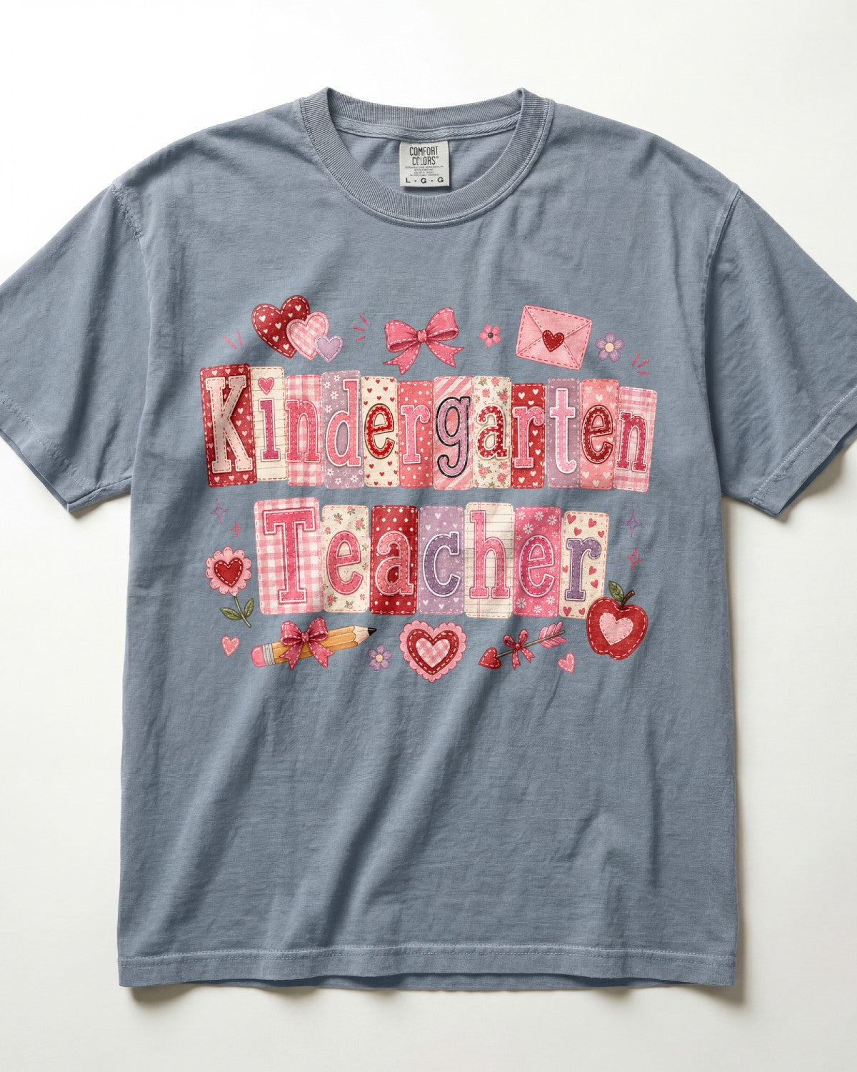 A slate blue/blue jean short-sleeve t-shirt with a 'Kindergarten Teacher' graphic on the front, featuring a patchwork-style design in pink, red, and white with hearts, bows, and strawberries, laid flat against a white background.