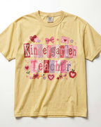 A pale yellow/butter short-sleeve t-shirt with a 'Kindergarten Teacher' graphic print in pink and red patchwork lettering, decorated with hearts, bows, and apples on a white background.